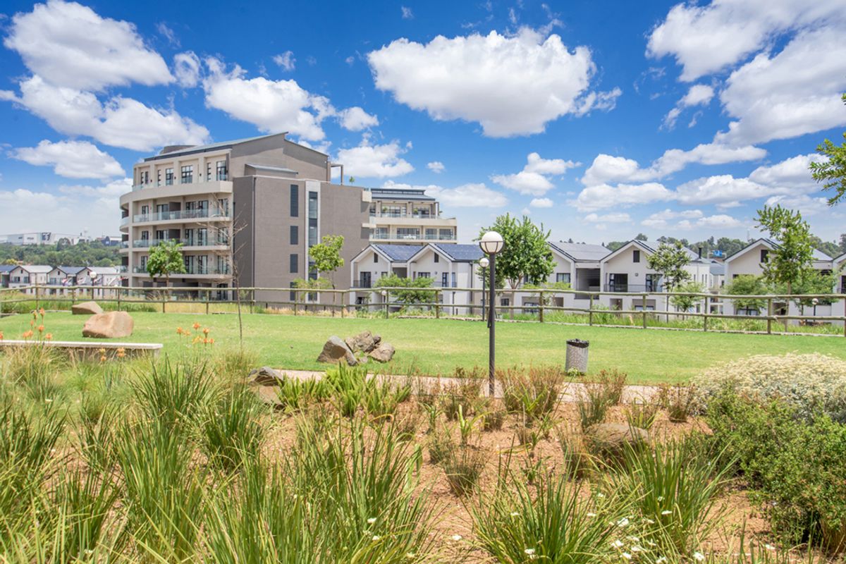 A well-maintained communal garden area featuring landscaped greenery, paved pathways, and timber fencing, set against a backdrop of modern apartment buildings.