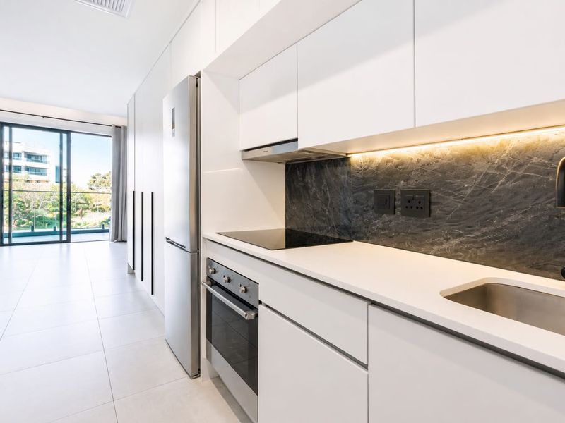Modern open-plan kitchen featuring sleek white cabinetry, stone countertops, integrated appliances, and a dark stone splashback, leading to a bright living area with balcony access.