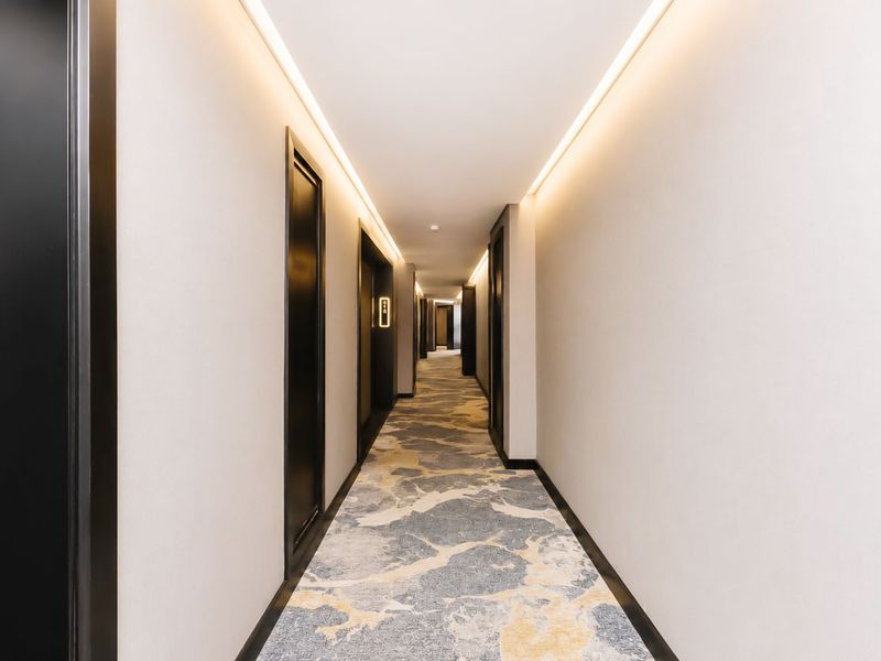 A modern, well-lit apartment hallway featuring clean white walls, black door frames, and patterned carpet flooring with integrated recessed cove lighting.