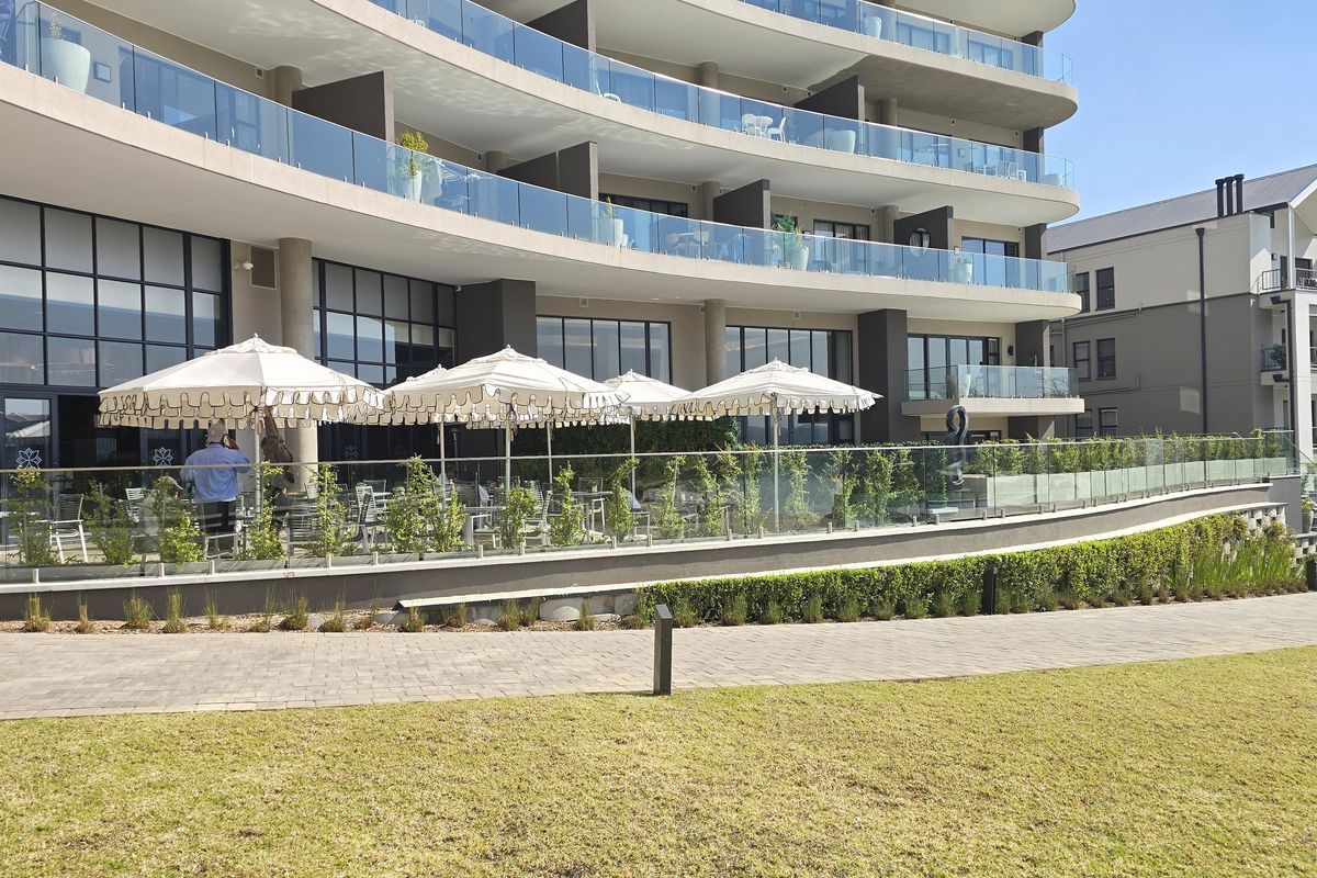 A modern apartment building exterior featuring multi-level glass balconies, large windows, and a well-maintained landscaped garden with a paved walkway.