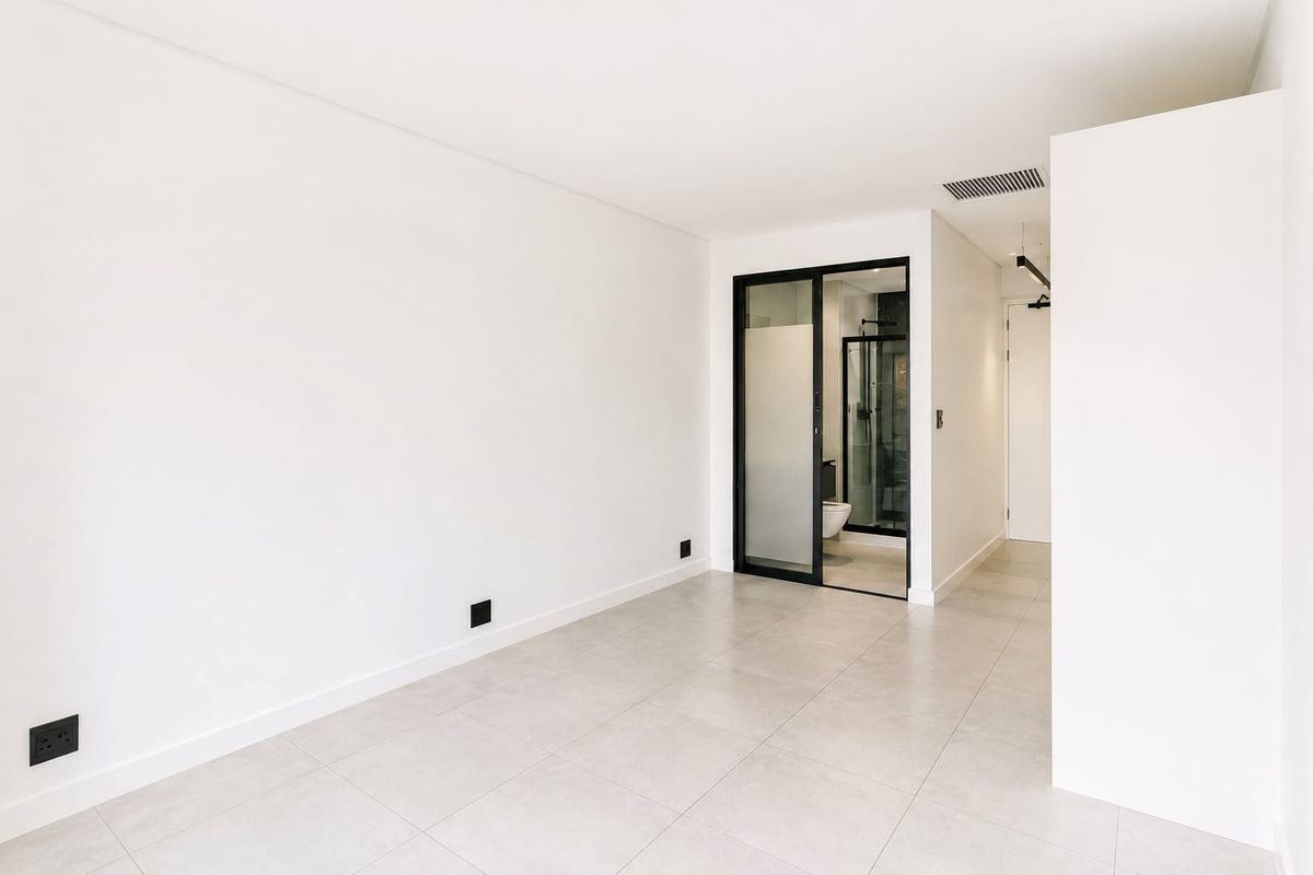 A bright, modern, and empty bedroom featuring neutral tiled flooring, clean white walls, and a sleek black-framed glass door leading to an en-suite bathroom.