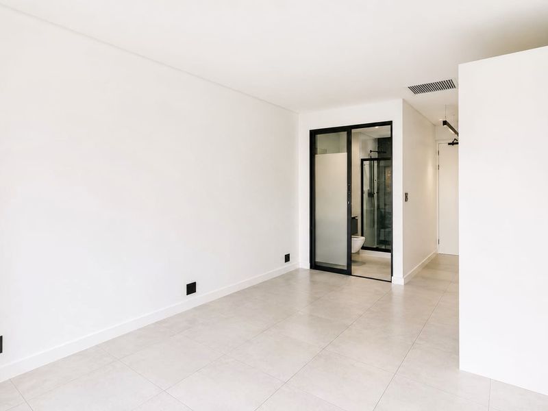 A bright, modern, and empty bedroom featuring neutral tiled flooring, clean white walls, and a sleek black-framed glass door leading to an en-suite bathroom.