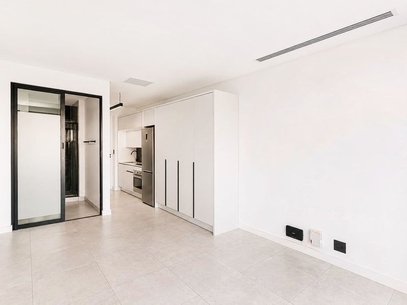 Modern open-plan living area featuring sleek white cabinetry, integrated kitchen appliances, tiled flooring, and ducted air conditioning in a bright, minimalist apartment setting.