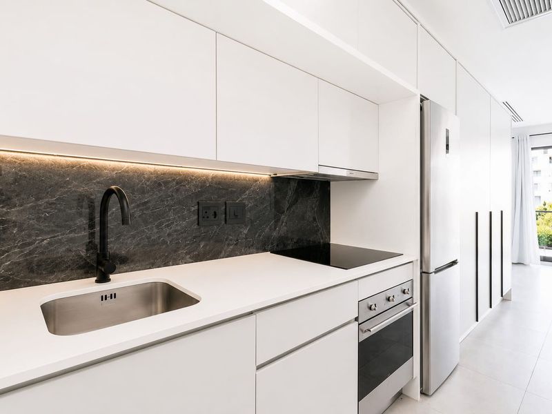 Modern kitchen featuring sleek white cabinetry, dark stone backsplash, integrated appliances, and high-quality finishes, leading to a bright balcony area.
