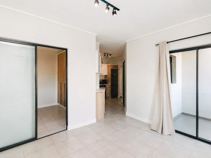 Bright open-plan living area featuring tiled floors, modern track lighting, and sliding glass doors leading to an outdoor space, with an adjacent kitchen area.