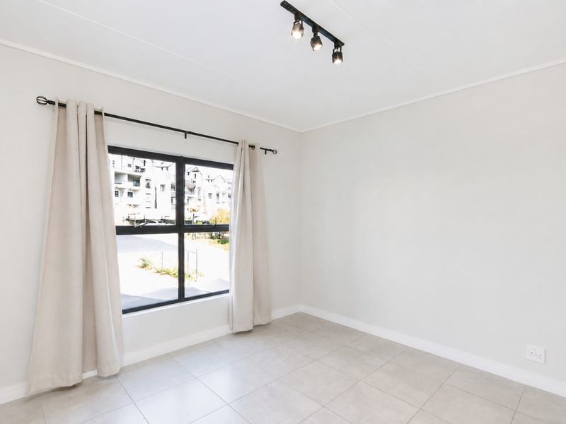 A bright, empty bedroom featuring neutral tiled flooring, modern track lighting, and a large window with black frames.