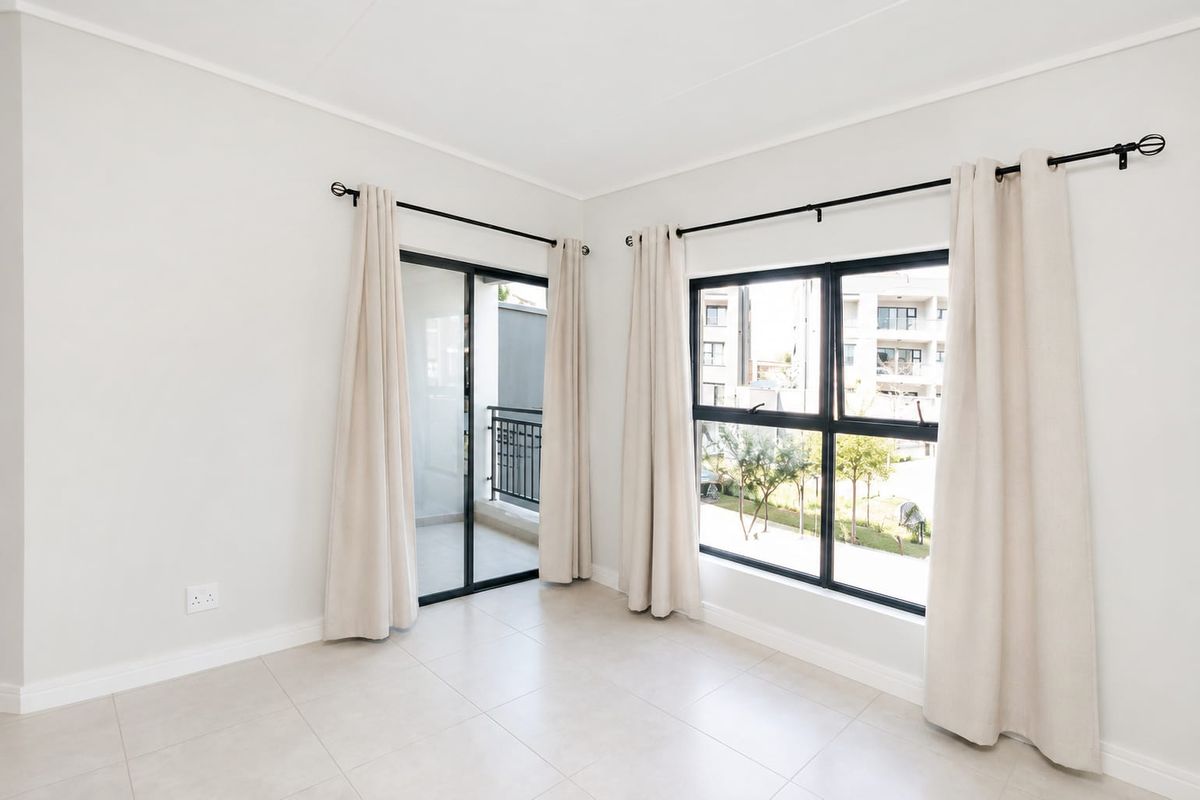 A bright, empty living area featuring neutral tiled flooring, large aluminium-framed windows, and a sliding door leading to a balcony.