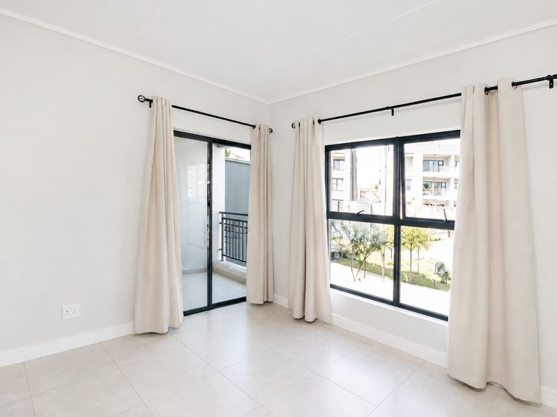 A bright, empty living area featuring neutral tiled flooring, large aluminium-framed windows, and a sliding door leading to a balcony.