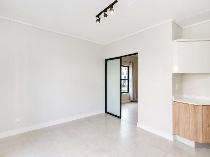 A bright, modern open-plan living area featuring neutral tiled floors, contemporary track lighting, and sleek built-in cabinetry with a stone countertop.