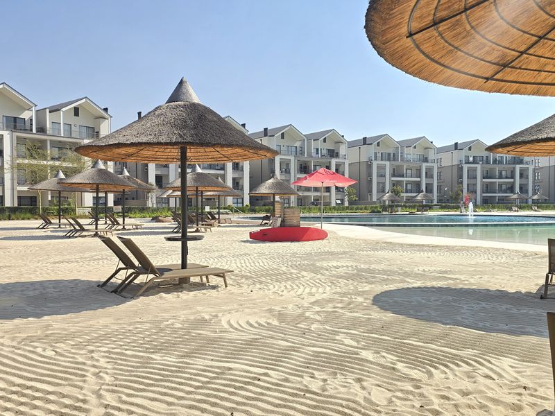 A well-maintained communal pool area featuring a sandy beach, thatched umbrellas, and modern apartment buildings in the background.
