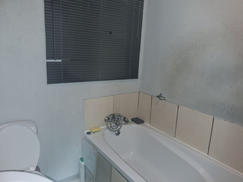 A bathroom featuring a bathtub with tiled splashback, toilet, and window blinds. The space requires cosmetic updates and wall repairs.