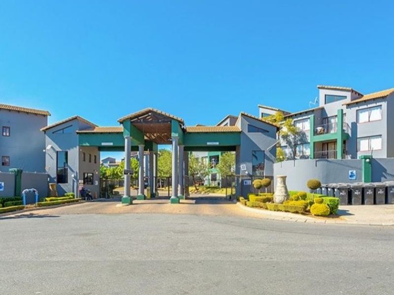 The main entrance and security gate of the apartment complex, featuring a paved driveway and established perimeter walling.