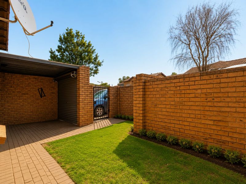 A well-maintained rear exterior area featuring a brick boundary wall, paved driveway, and a covered carport with a small lawn.