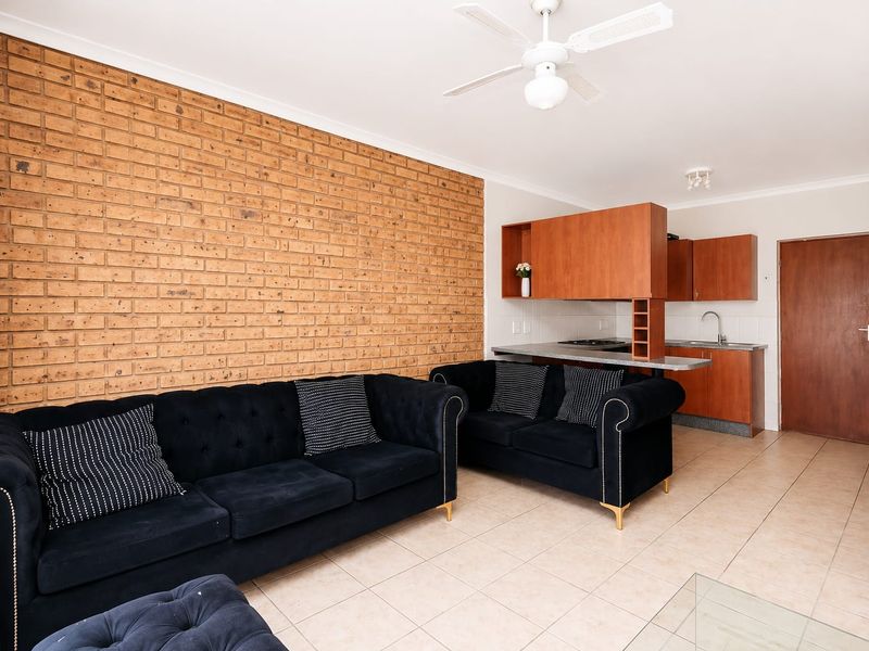 Open-plan living area featuring an exposed brick wall, tiled flooring, ceiling fan, and a functional kitchen with built-in cabinetry and a breakfast bar.