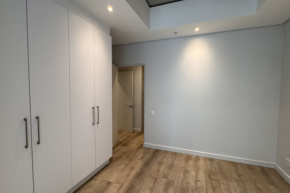 A clean, modern bedroom featuring neutral walls, light wood-look laminate flooring, and ample built-in cupboard storage with recessed ceiling lighting.