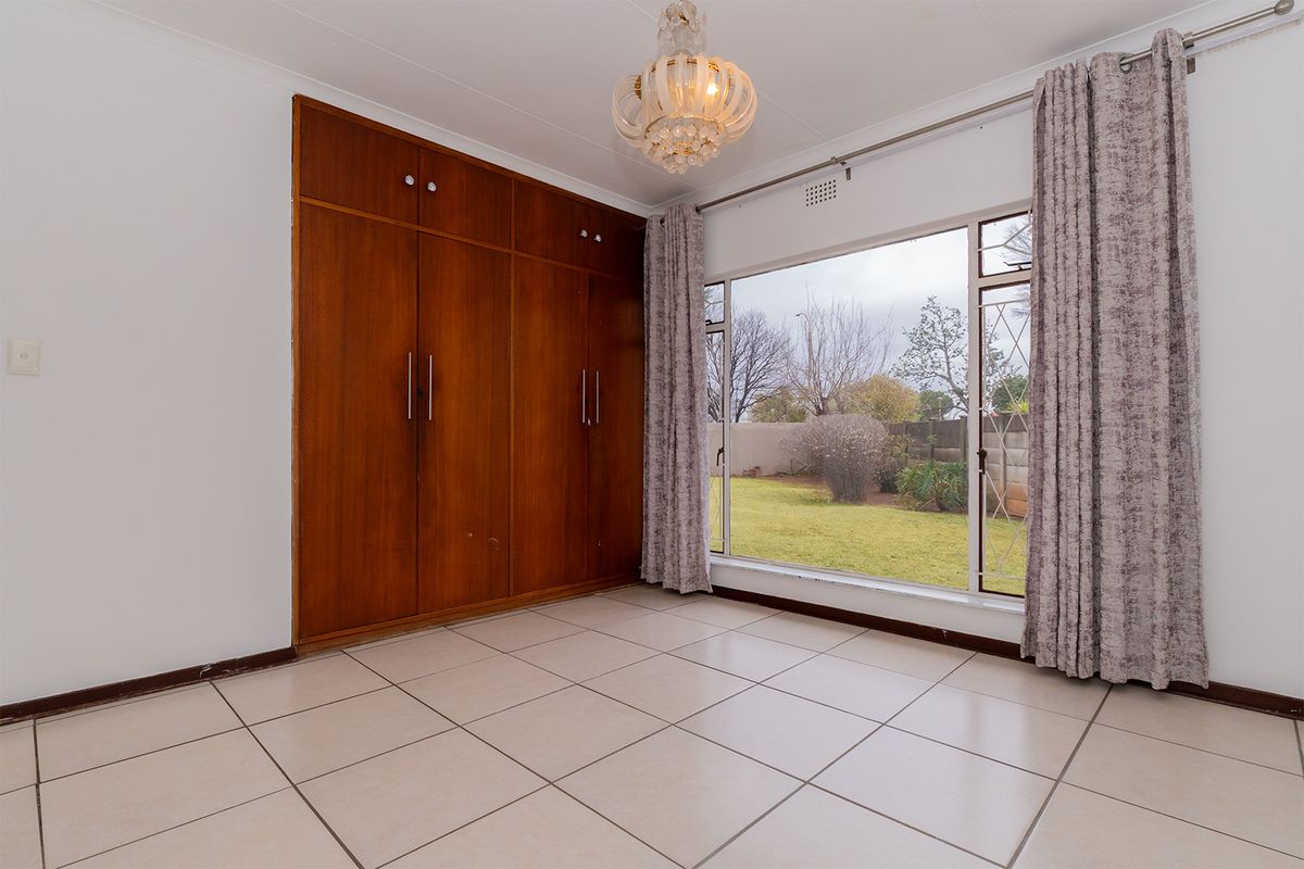 A bright, empty bedroom featuring tiled flooring, large windows with garden views, and ample built-in wooden cupboard storage.