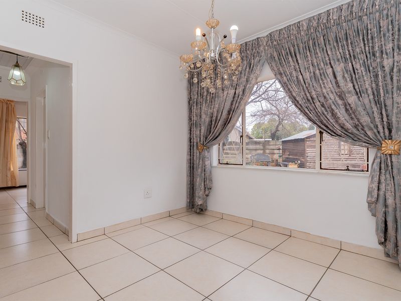 A bright, empty dining room featuring neutral tiled flooring, a decorative chandelier, and large windows with security bars overlooking the rear yard.
