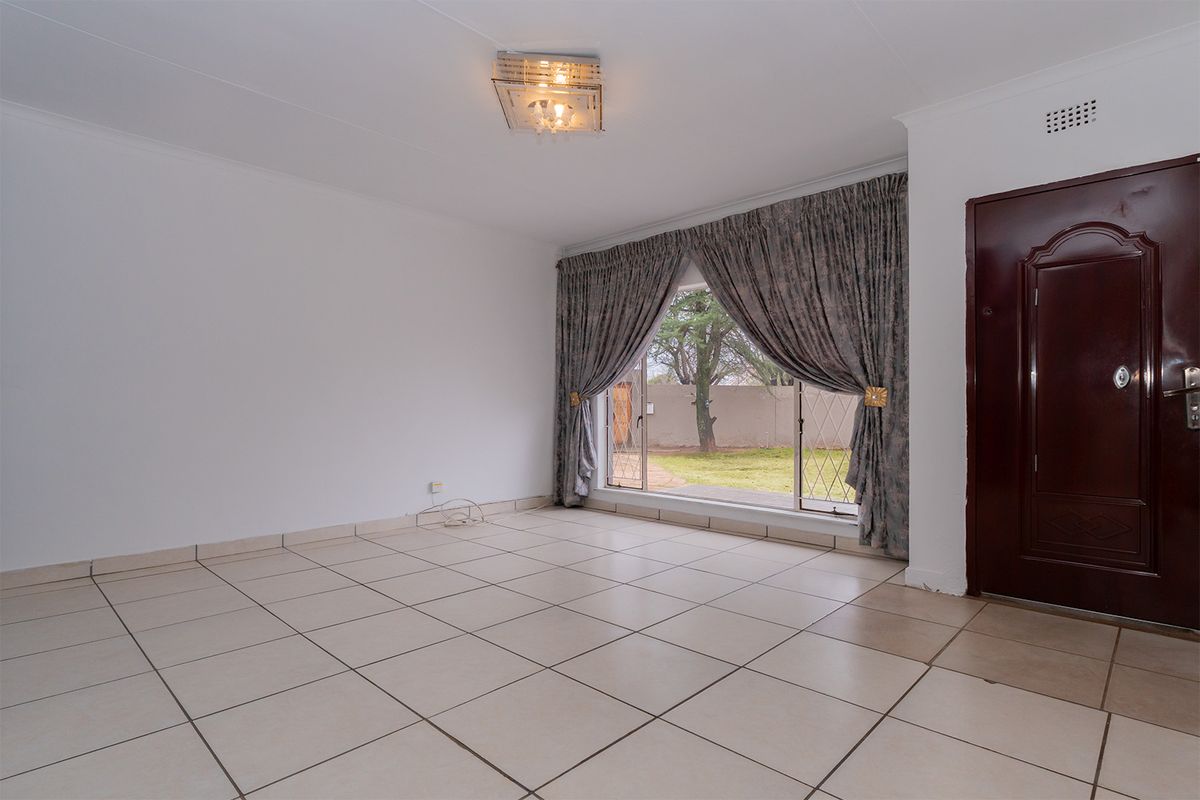 A bright, empty living room featuring neutral tiled flooring, a large sliding glass door with garden access, and a decorative ceiling light fixture.