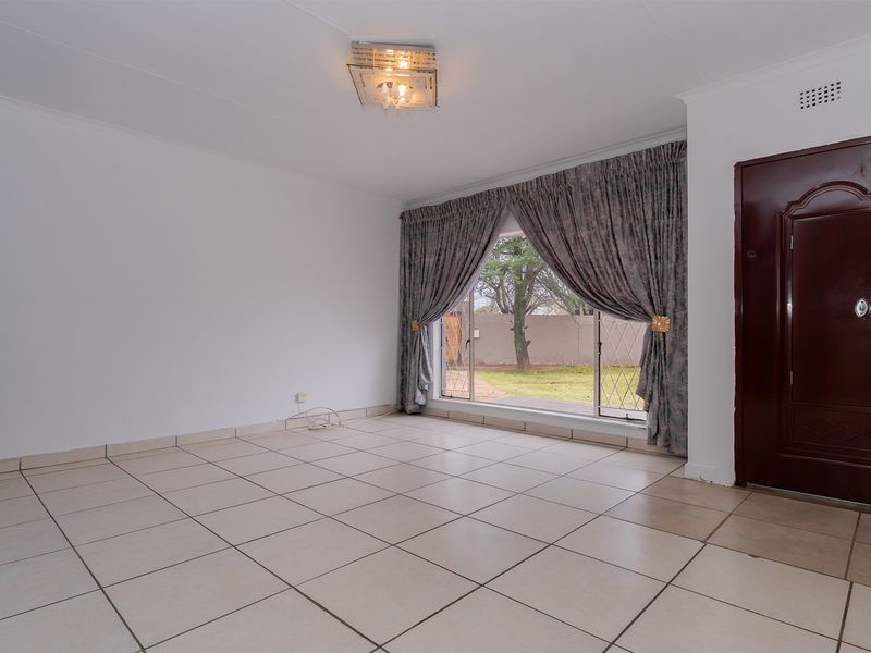 A bright, empty living room featuring neutral tiled flooring, a large sliding glass door with garden access, and a decorative ceiling light fixture.