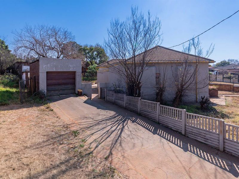 Front view of the property featuring a concrete driveway, single garage, and perimeter boundary wall, set on a large plot with established trees.