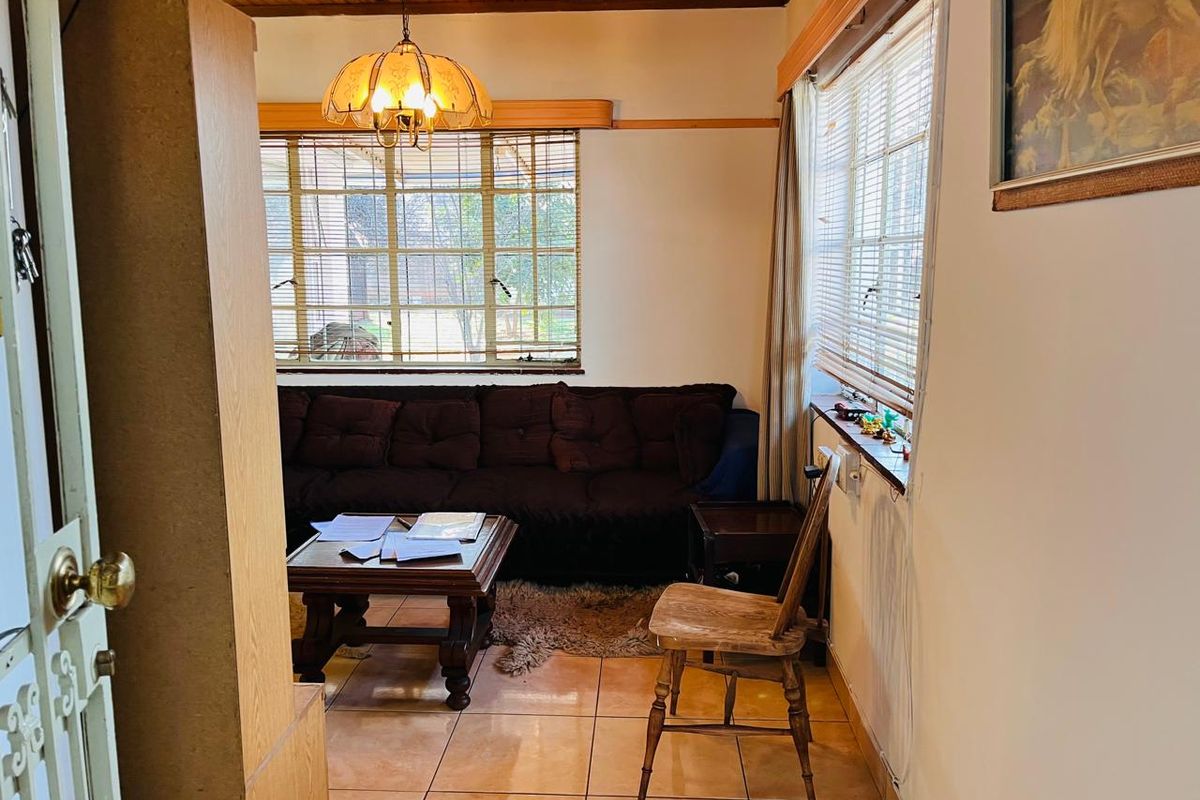 A living room featuring ceramic floor tiles, timber ceiling paneling, and large windows with venetian blinds, offering a dated but functional living space.
