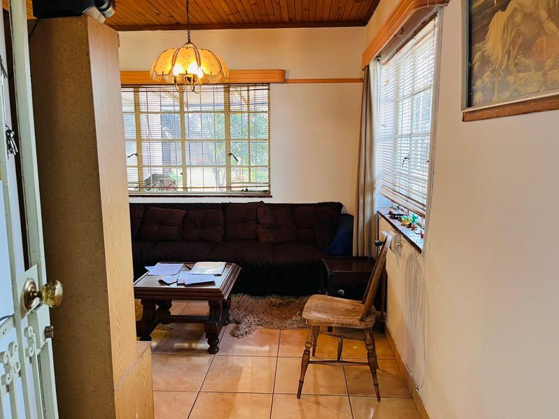 A living room featuring ceramic floor tiles, timber ceiling paneling, and large windows with venetian blinds, offering a dated but functional living space.