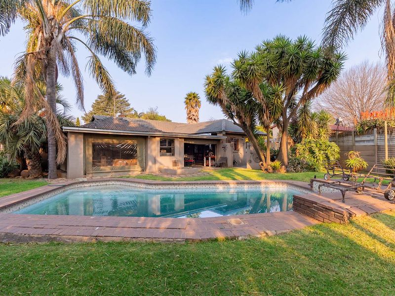 A rear garden view featuring a swimming pool with brick coping, mature trees, and a single-story house with a tiled roof.