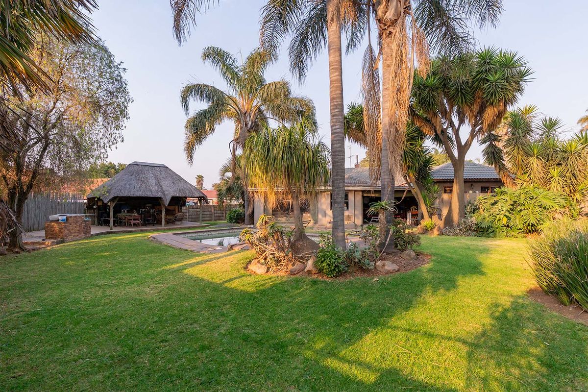 Spacious rear garden featuring a swimming pool, mature palm trees, and a traditional thatched lapa with a built-in braai area.