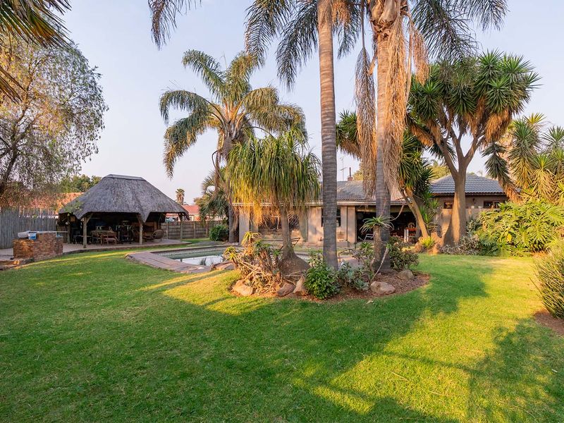 Spacious rear garden featuring a swimming pool, mature palm trees, and a traditional thatched lapa with a built-in braai area.