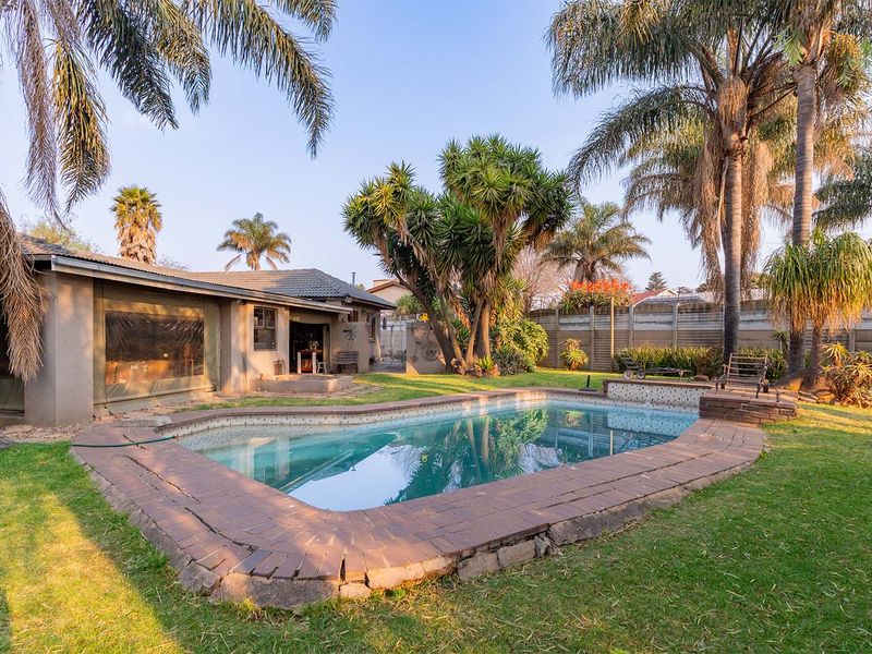 A backyard area featuring a swimming pool with brick coping, surrounded by a lawn, mature palm trees, and a precast concrete boundary wall.