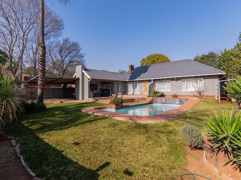 Rear view of the property featuring a swimming pool, covered patio area, carport, and a well-maintained garden with mature trees.