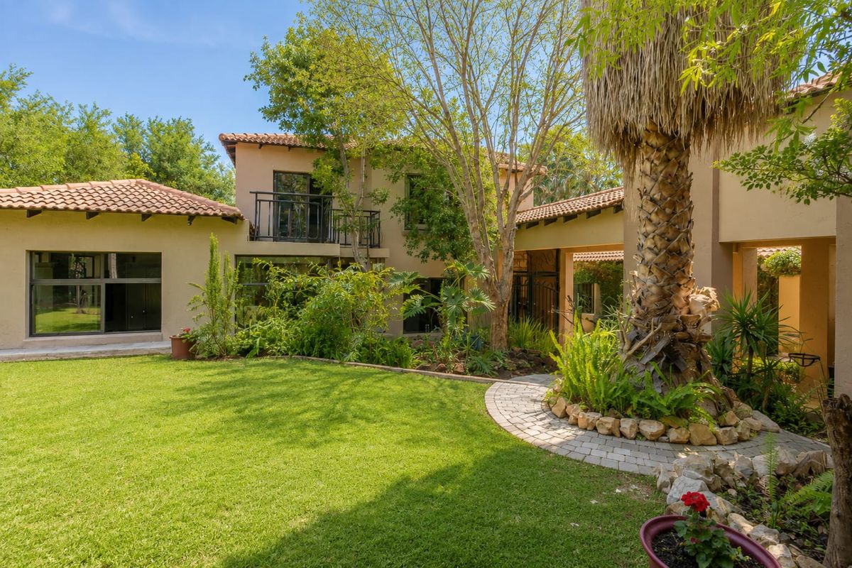 A view of the rear exterior of the house featuring a landscaped garden, mature trees, a paver path, and a balcony with metal railings.