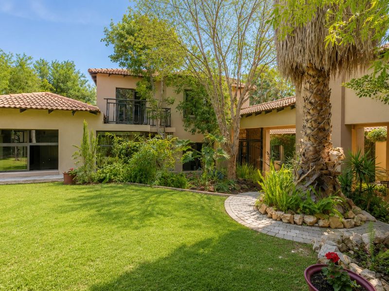 A view of the rear exterior of the house featuring a landscaped garden, mature trees, a paver path, and a balcony with metal railings.
