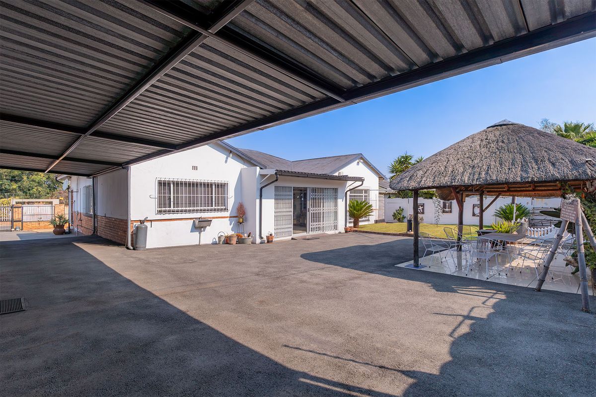 Rear exterior view featuring a large paved driveway, covered carport, and a thatched lapa entertainment area set within a private garden space.