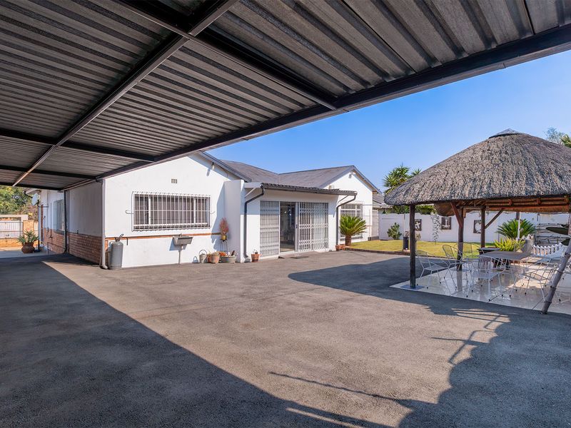 Rear exterior view featuring a large paved driveway, covered carport, and a thatched lapa entertainment area set within a private garden space.