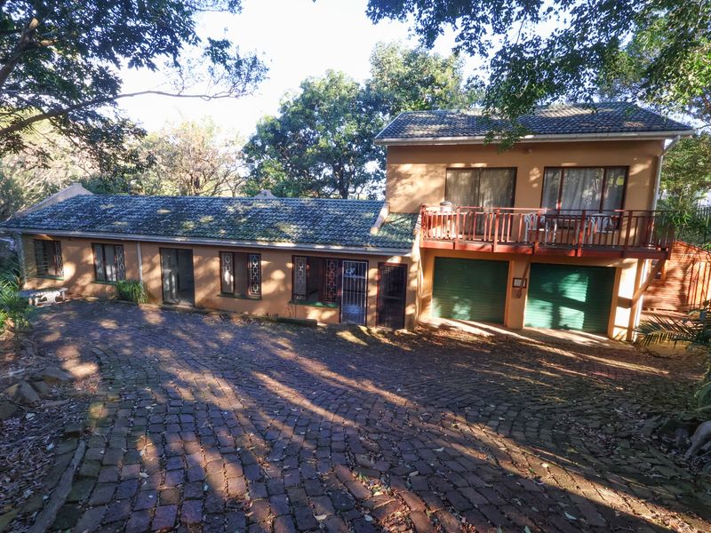 Front elevation of a two-story house featuring a cobblestone driveway, double garage, and an upper-level balcony surrounded by mature trees.