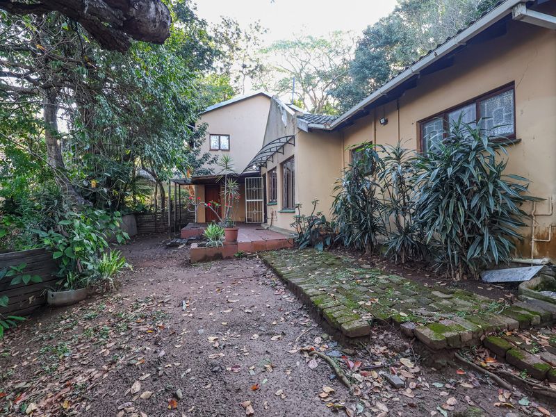 Rear exterior view of the house featuring a tiled patio, brick pathway, and established garden area requiring maintenance.