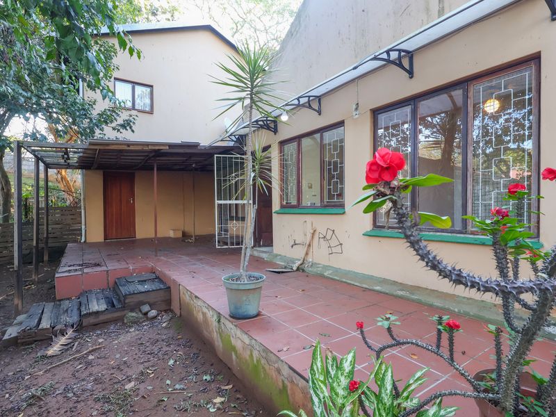 Rear exterior view featuring a tiled patio area, metal awning, and security features on windows and doors, showing signs of weathering.