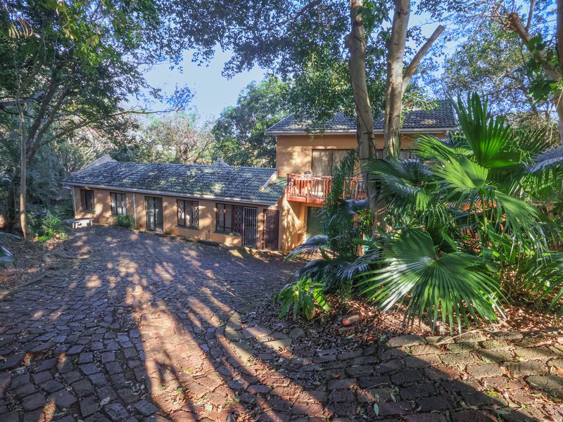 A multi-level house exterior featuring a cobblestone driveway, mature trees, and a balcony, set within a lush, wooded environment.