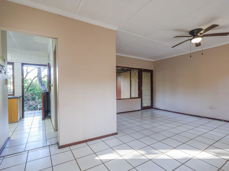An empty, open-plan living area featuring tiled flooring, a ceiling fan, and direct access to an adjacent kitchen and outdoor space.