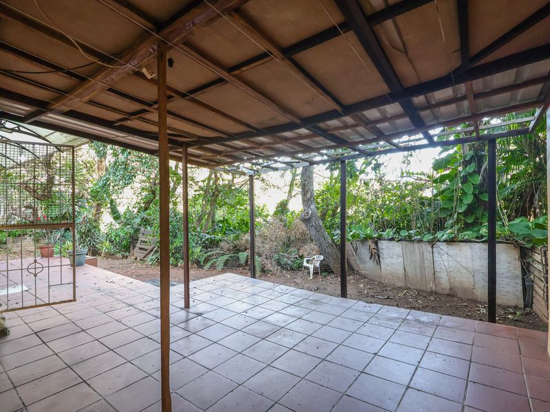 A covered outdoor patio area featuring tiled flooring and metal support structures, overlooking a garden space with a retaining wall.