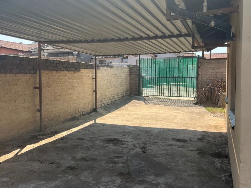 A rear exterior area featuring a covered carport structure with a corrugated metal roof, concrete paving, and a secure metal gate.