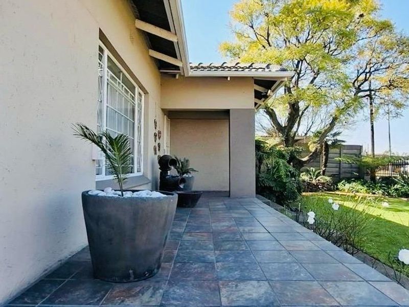 A covered patio area featuring tiled flooring, overlooking a well-maintained garden with a wrought iron entrance gate.