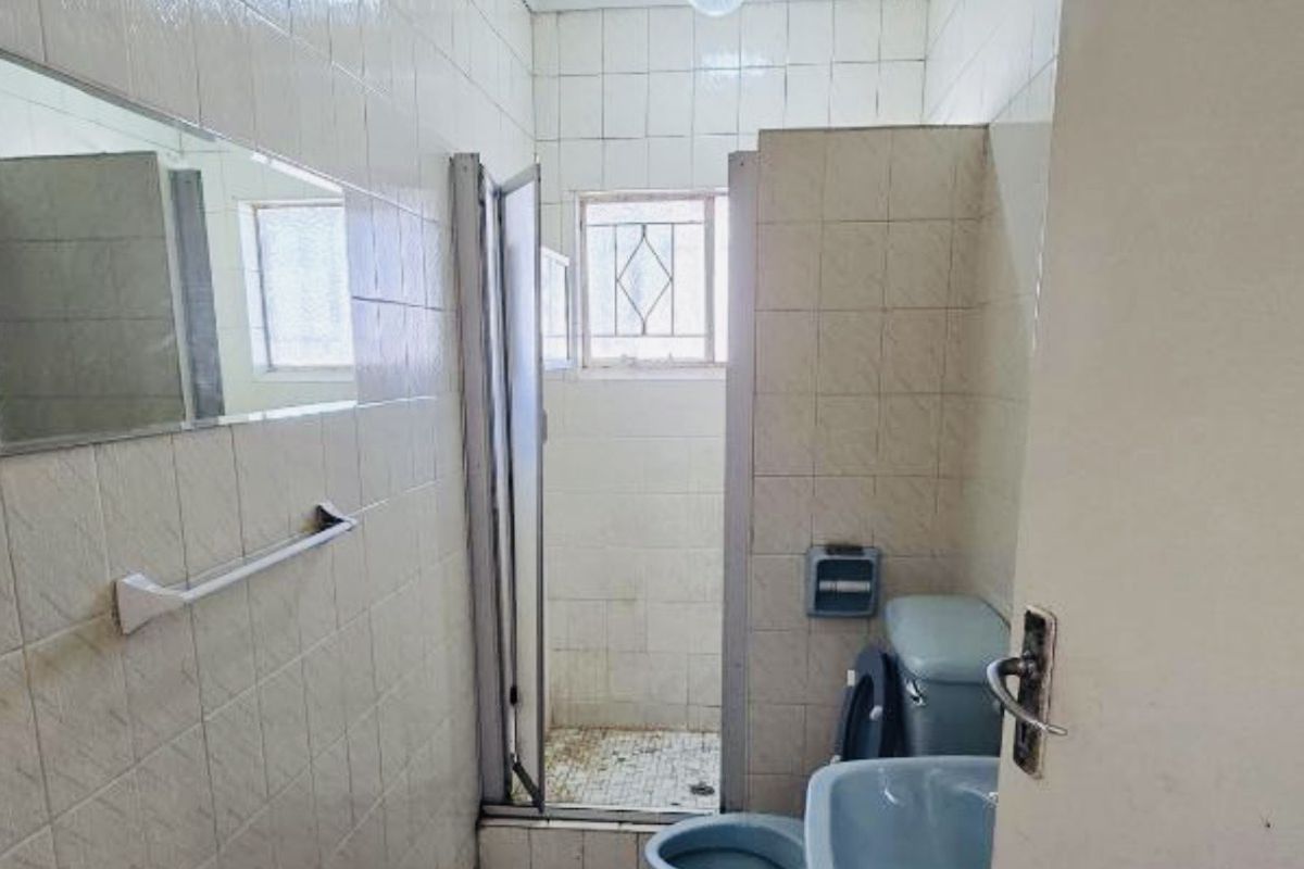 A dated bathroom featuring tiled walls and floors, a pedestal basin, toilet, and a shower enclosure with visible signs of wear and ceiling staining.