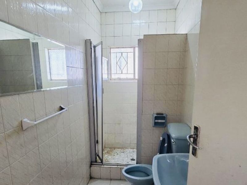 A dated bathroom featuring tiled walls and floors, a pedestal basin, toilet, and a shower enclosure with visible signs of wear and ceiling staining.