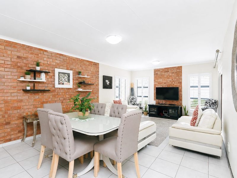 Spacious open-plan living and dining area featuring an exposed brick feature wall, neutral tiled flooring, and plantation shutters for natural light control.