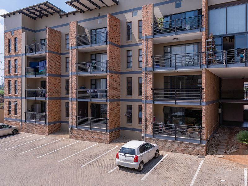 Exterior view of a multi-story apartment complex featuring brick facade detailing, private balconies with metal railings, and a paved ground-level parking area.