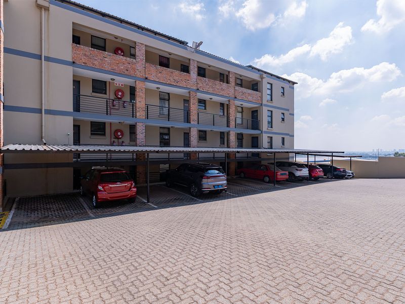 Exterior view of a multi-story apartment complex featuring a paved parking area with covered bays and brick facade detailing.