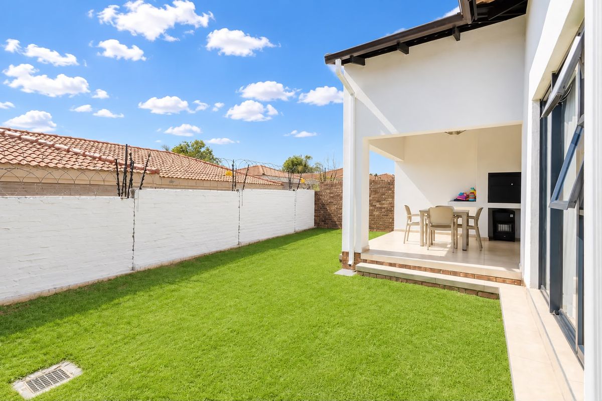 A well-maintained rear garden featuring a covered patio with a built-in braai, tiled flooring, and a secure brick boundary wall.