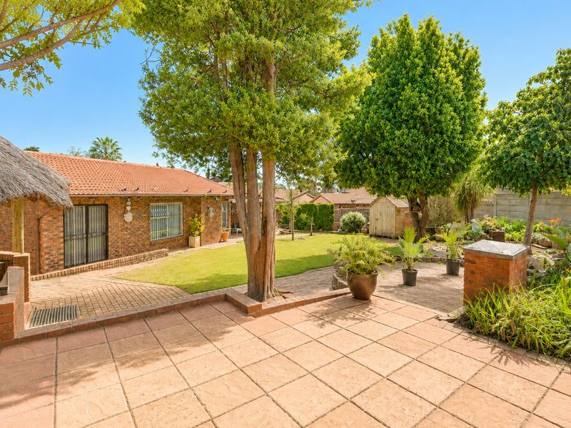 A spacious rear garden featuring a paved patio area, a thatched lapa, established trees, and a well-maintained lawn with a brick-built house facade.